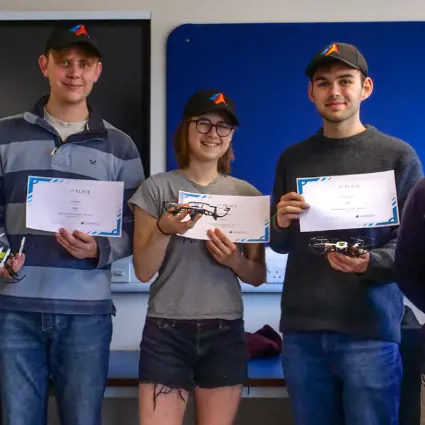 A full team photo of Team Orville who competed in the MathWorks Minidrone Competition. There are four men and one woman, with two men standing either side of the woman. The three people in the middle are holding the award certificates and drones they used.