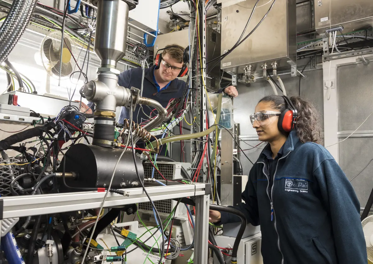 Two students with ear protectors and protective eye glasses look at a big complicated machine