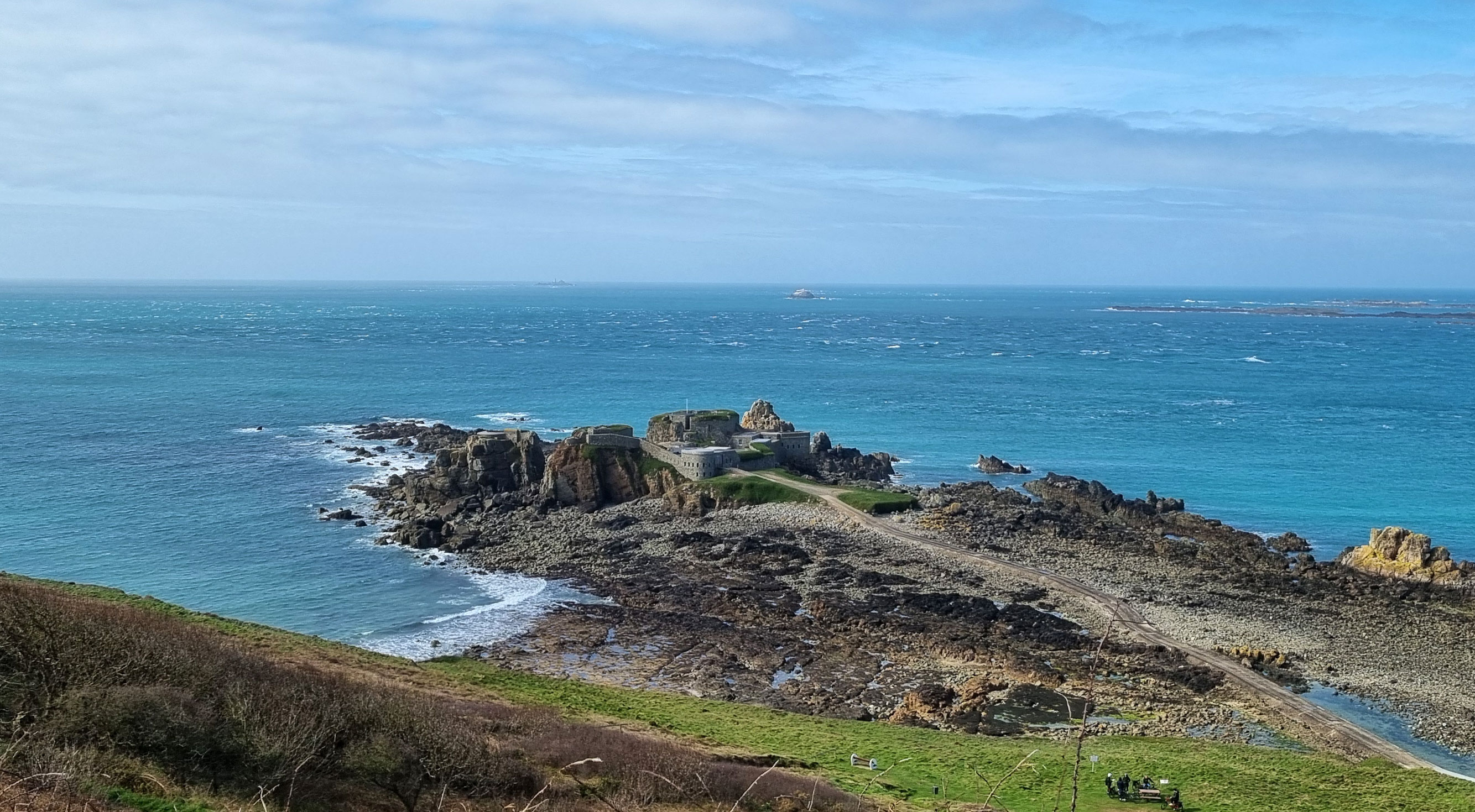 A coastal view of Fort Clonque on Alderney, connected to the mainland by a narrow causeway, surrounded by rocky shoreline and blue sea.