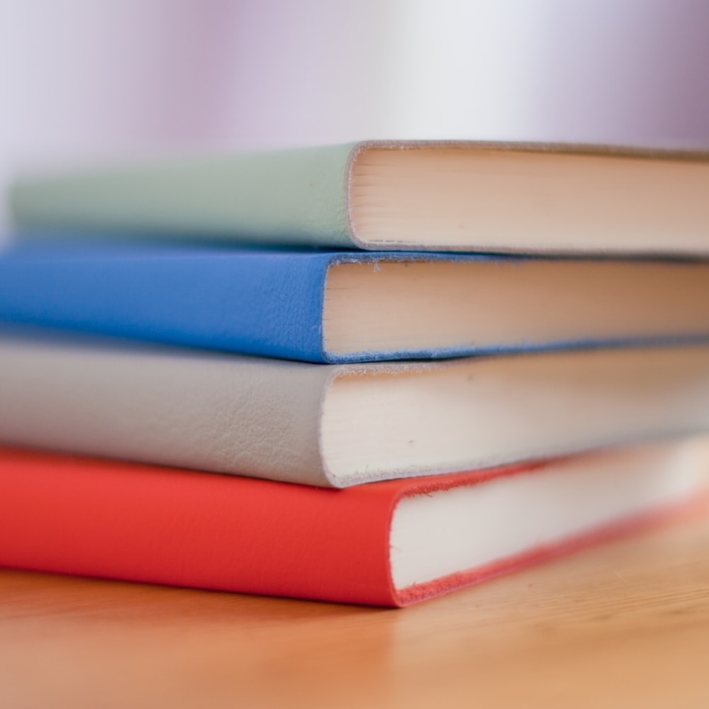 A pile of 4 books in red, grey, blue and light green, with their spines facing the camera