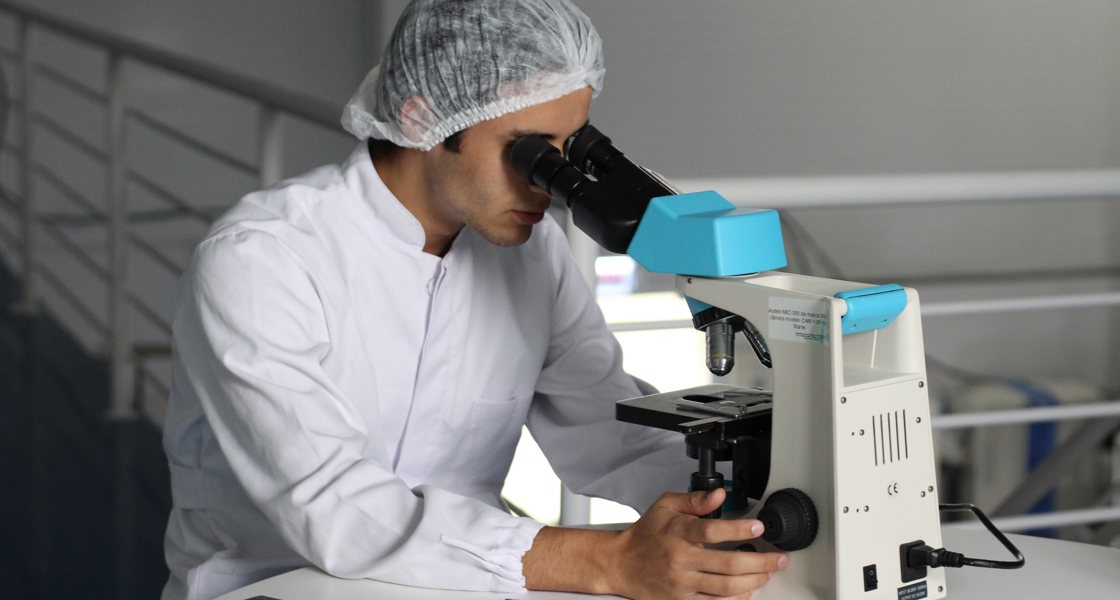 Someone in a white jacket and hairnet looking into a microscope
