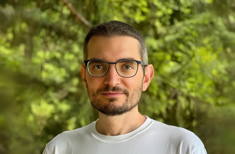 Portrait of Professor Andrea Vedaldi wearing glasses and a white shirt, standing outdoors with a background of green trees and soft natural light.