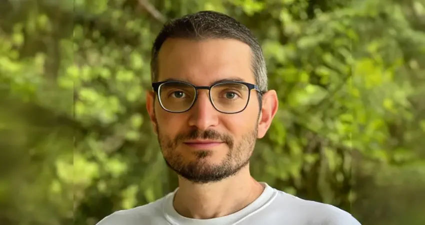 Portrait of Professor Andrea Vedaldi wearing glasses and a white shirt, standing outdoors with a background of green trees and soft natural light.