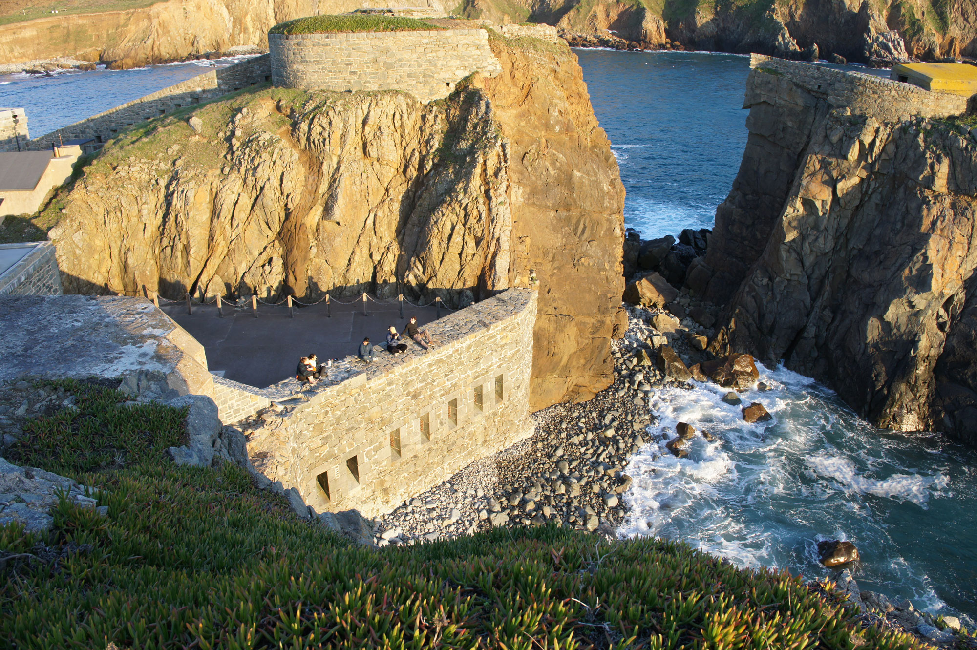 Group members seated along a stone wall watching the sunset over the sea, with waves breaking against the rocks.