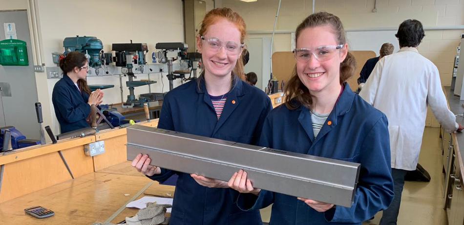 Two students in labcoats holding heavy bar