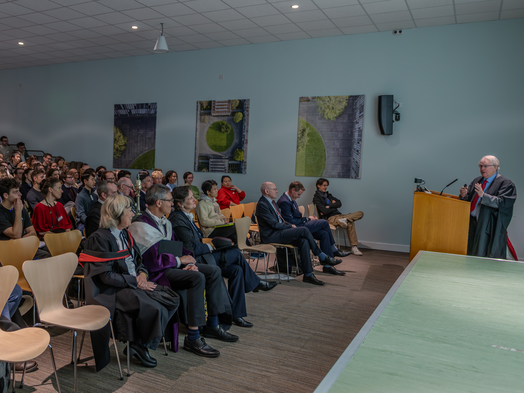Professor Stan Whittingham stands at a lectern delivering the John Goodenough Lecture in the Riverside Lecture Theatre at St Catherine’s College, speaking to a full audience seated across the room.