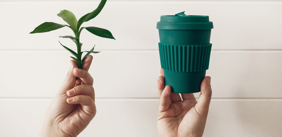 Hands holding stylish reusable eco coffee cup and green bamboo leaves on white wooden background
