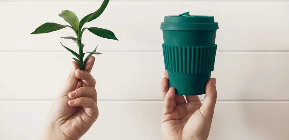 Hands holding stylish reusable eco coffee cup and green bamboo leaves on white wooden background