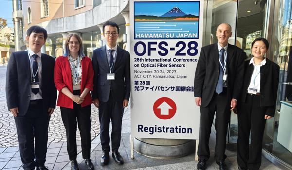 Oxford Team: five people are stood by a conference sign outside of a building. Three people stand on the left of the sign and two on the right, all are wearing business clothes suitable for a conference. On the left, we have an East Asian man, a white woman, and another East Asian man. Then on the right, we have a white man and an Asian woman.