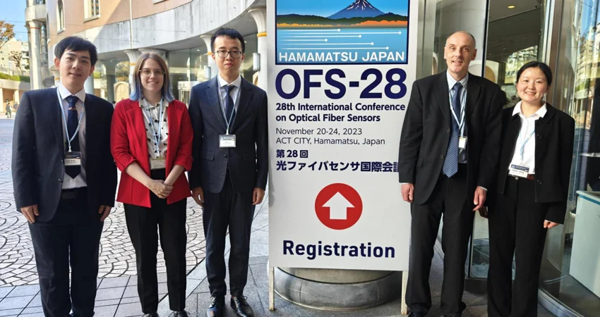 Oxford Team: five people are stood by a conference sign outside of a building. Three people stand on the left of the sign and two on the right, all are wearing business clothes suitable for a conference. On the left, we have an East Asian man, a white woman, and another East Asian man. Then on the right, we have a white man and an Asian woman.