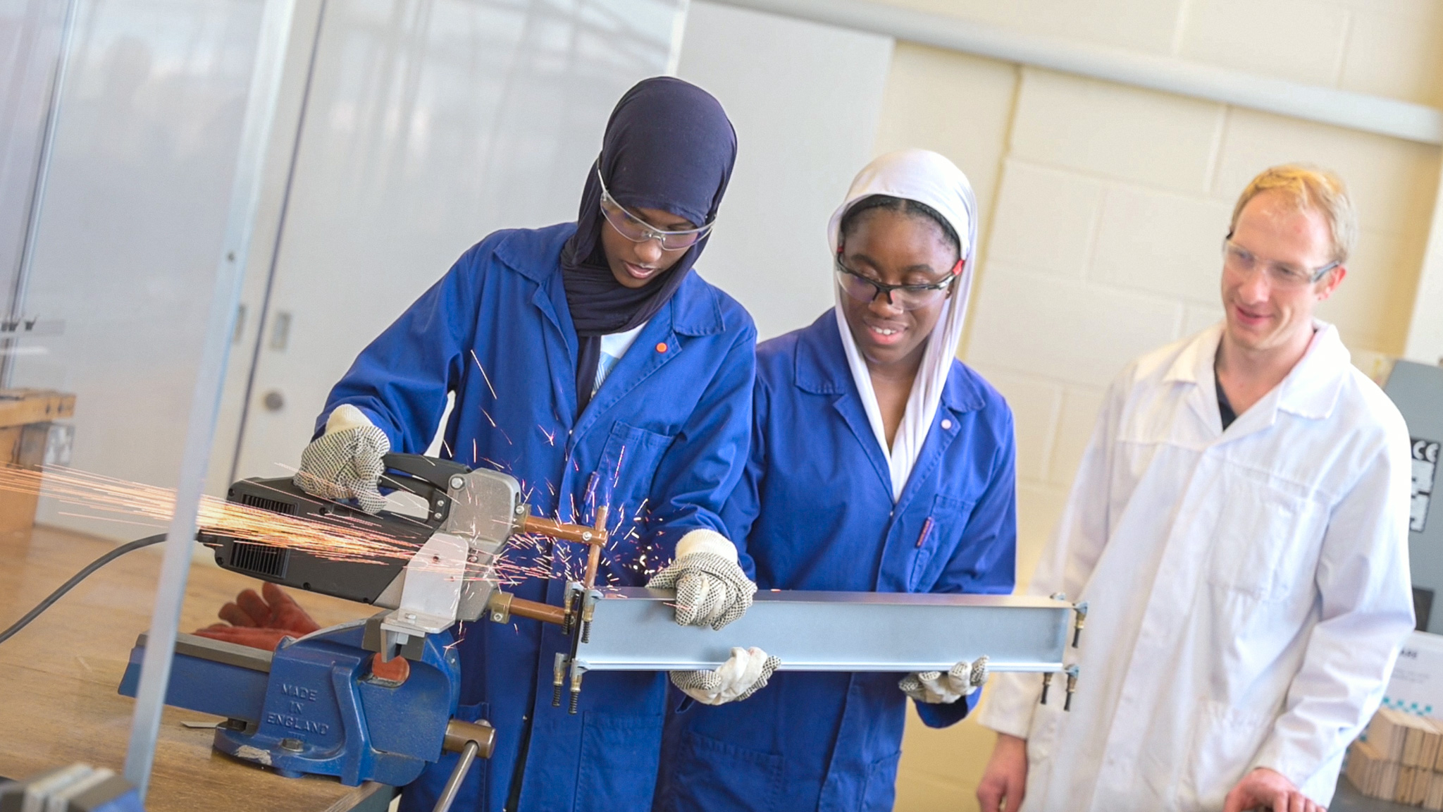 Two students wearing blue lab coats and safety goggles use a metal cutting tool during a hands-on engineering workshop while an instructor supervises.