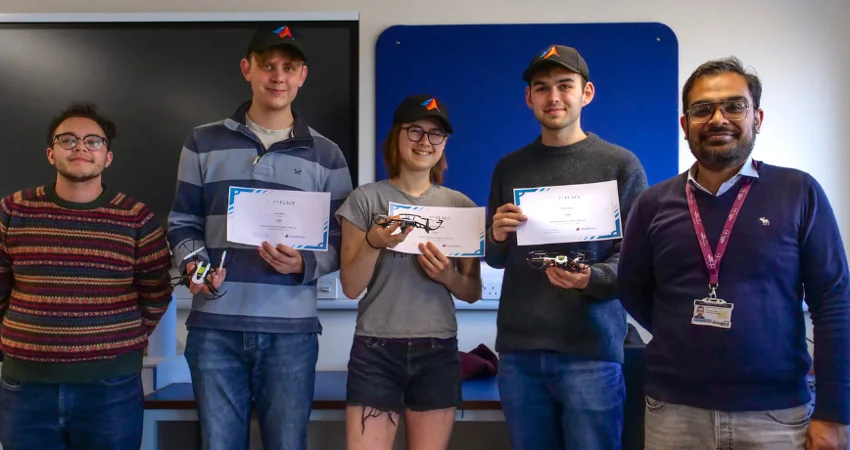 A full team photo of Team Orville who competed in the MathWorks Minidrone Competition. There are four men and one woman, with two men standing either side of the woman. The three people in the middle are holding the award certificates and drones they used.