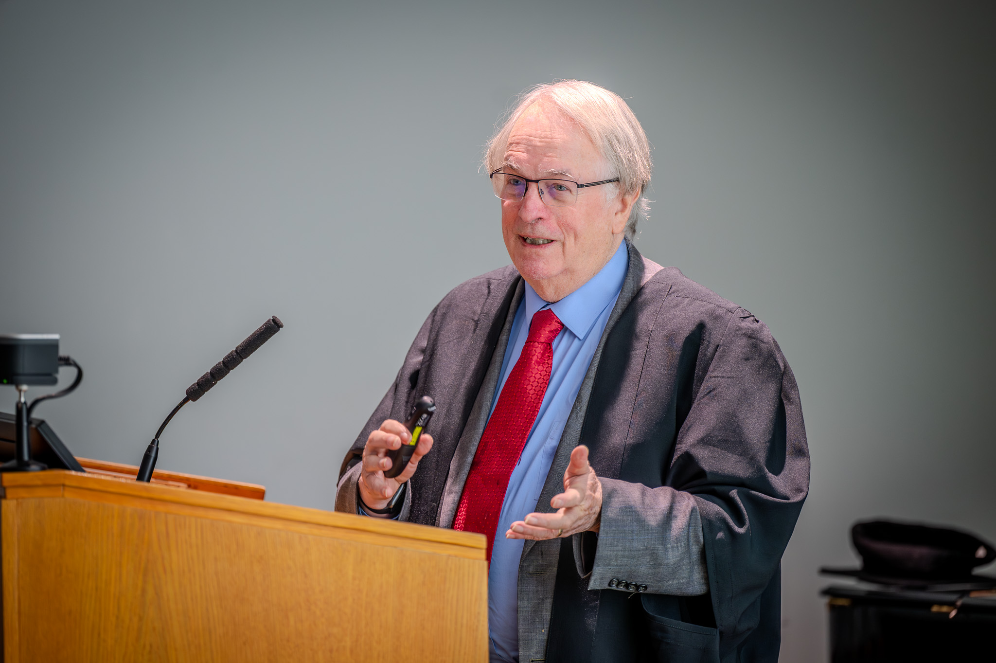 Professor Stan Whittingham stands at a wooden lectern delivering a lecture, smiling as he speaks to an audience. He is wearing academic robes and glasses, with microphones and screens in front of him in a bright lecture hall.