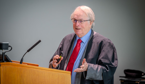 Professor Stan Whittingham stands at a wooden lectern delivering a lecture, smiling as he speaks to an audience. He is wearing academic robes and glasses, with microphones and screens in front of him in a bright lecture hall.
