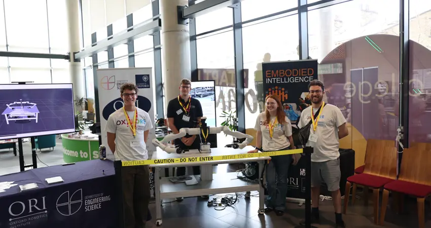 Four Oxford Robotics Institute team members pose with Frank the robot at the Oxplore Festival stand. Banners and educational materials promoting robotics research are visible behind them.