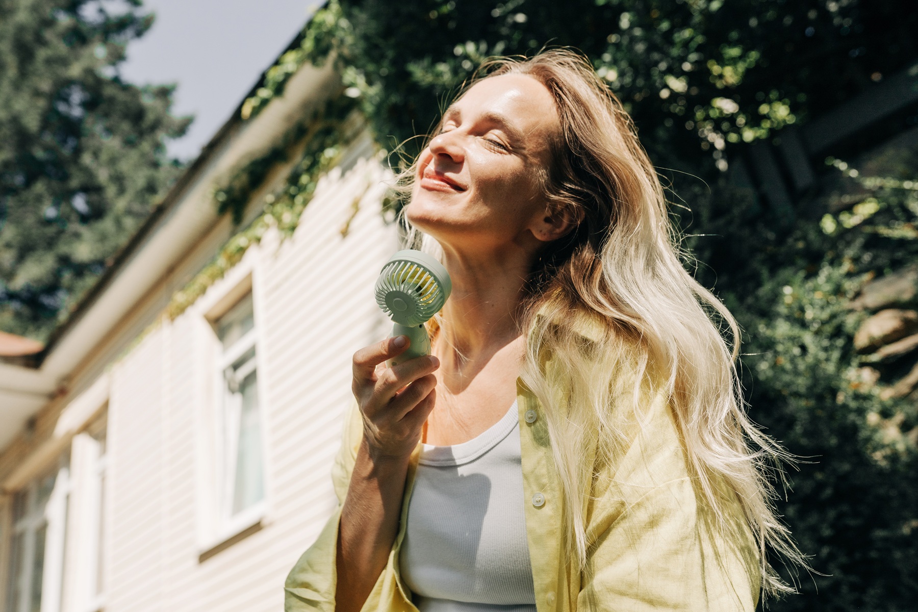 A happy smiling blonde woman uses a portable electric fan outdoors on a hot midday. By Ilona