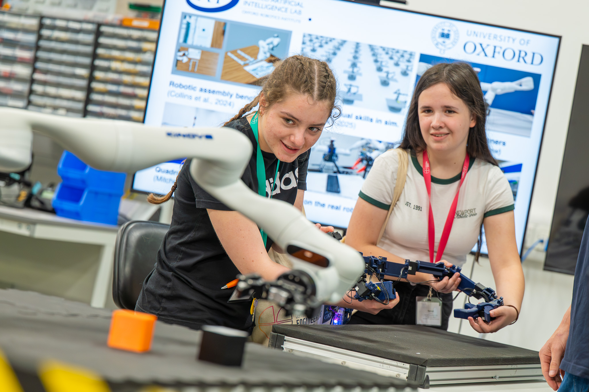 Two students take part in a robotics activity, interacting with a robotic arm in a lab while engineering demonstrations are displayed on screens behind them.