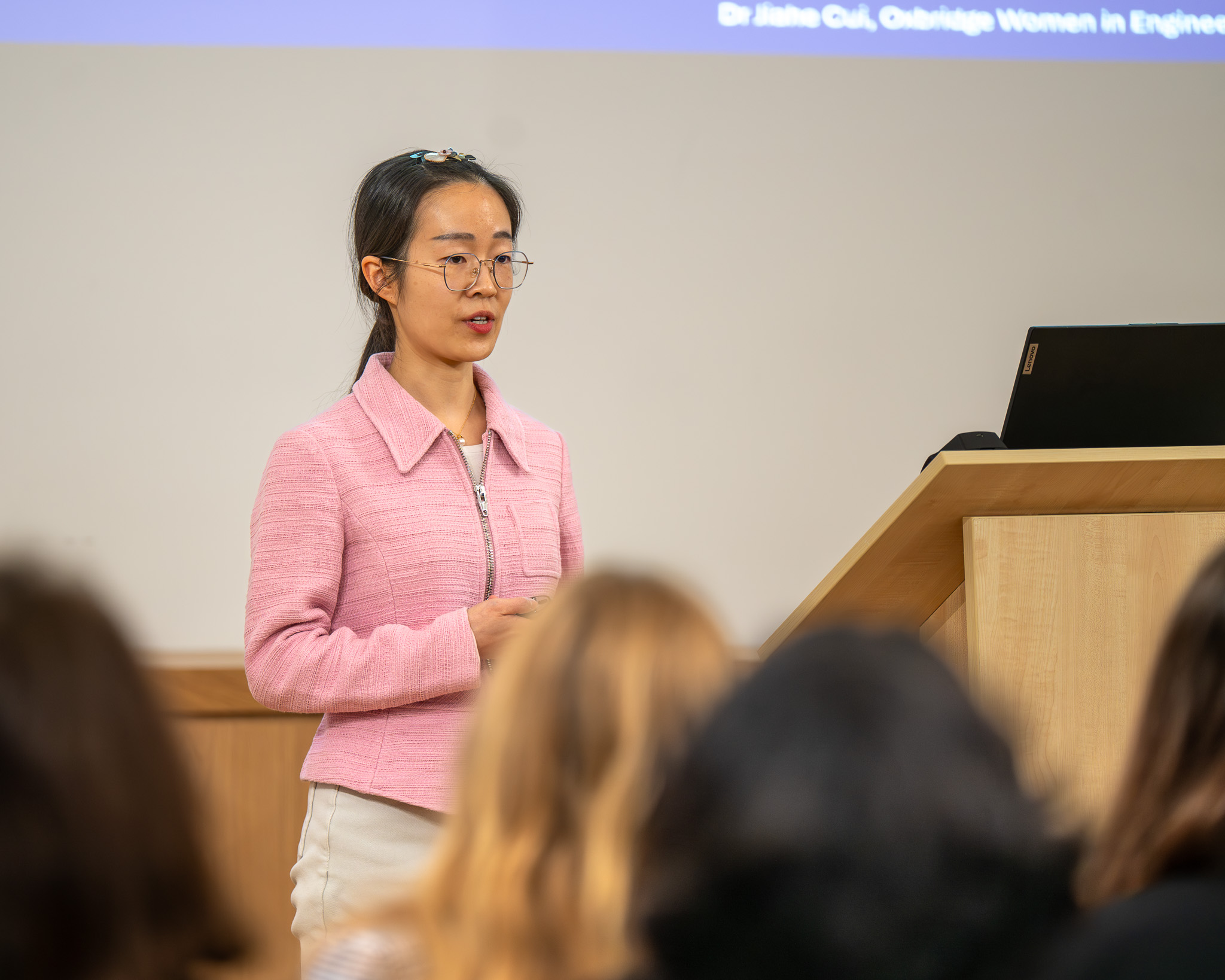 Dr Jiahe Cui stands at a lectern presenting to the audience, with part of her presentation slide visible behind her. She is speaking as part of the OxBridge Women in Engineering event.