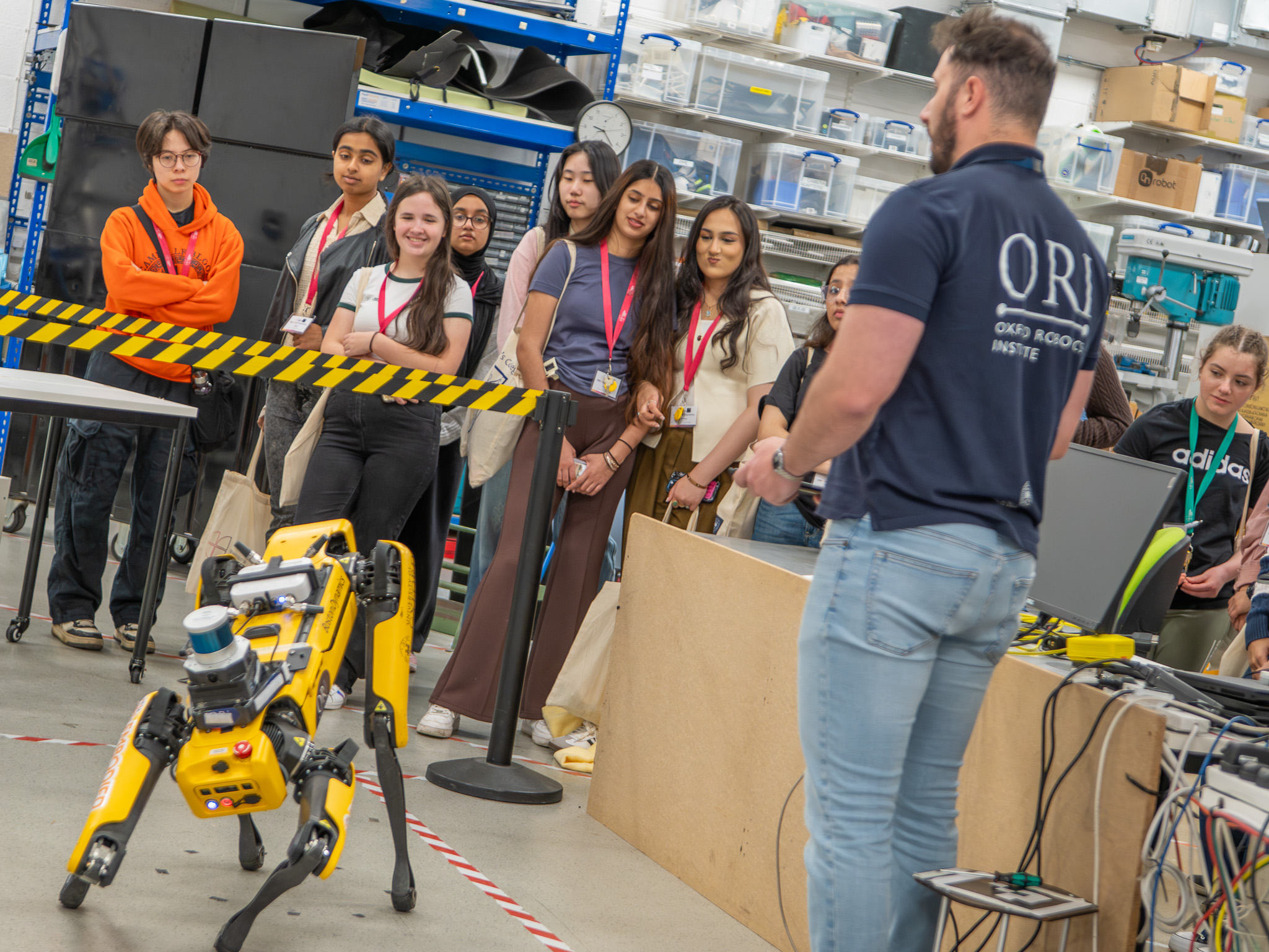 Students watch a demonstration from a member of the Oxford Robotics Institute as a yellow robotic dog moves across the lab floor.