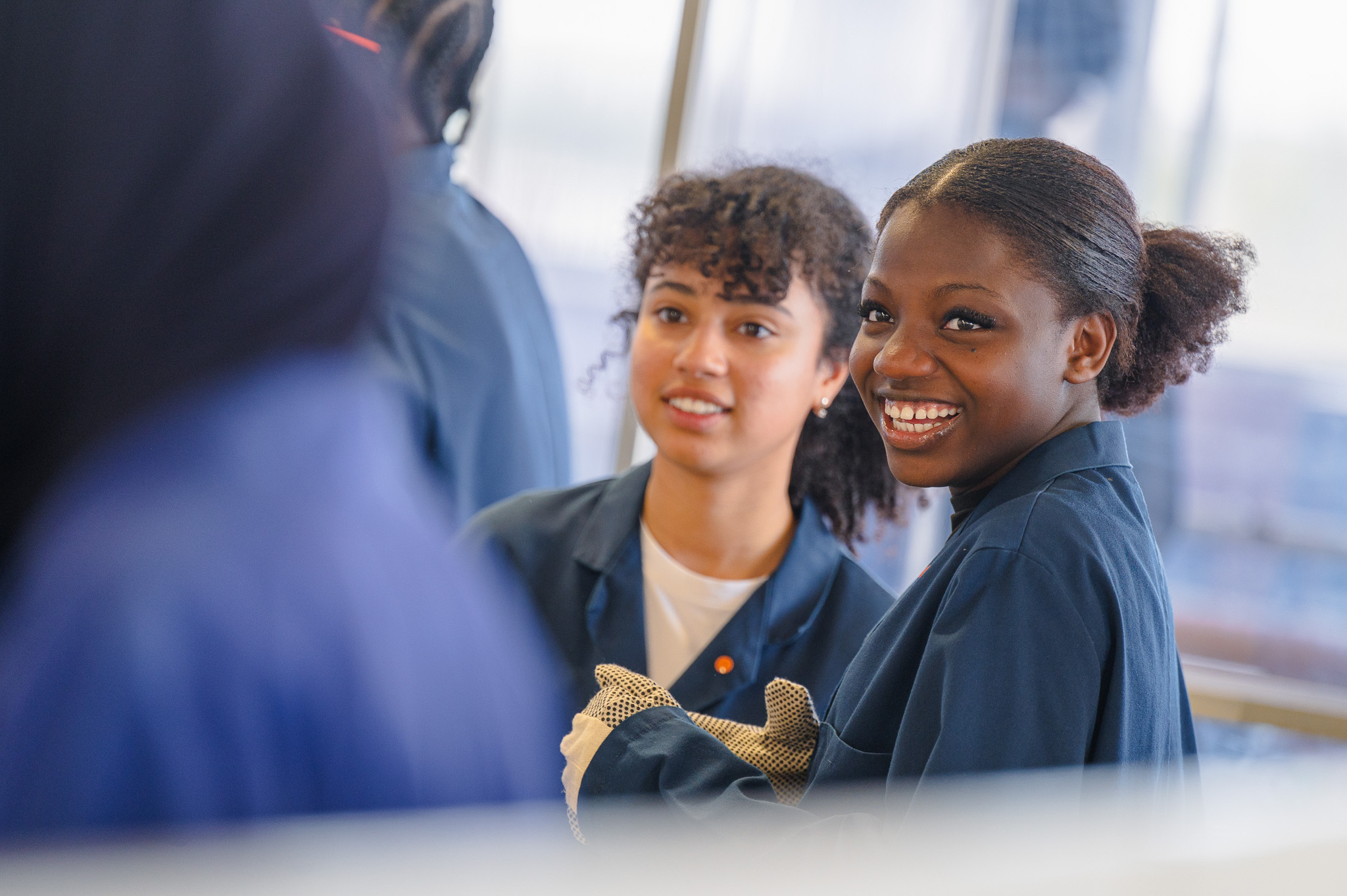 Two students in workshop overalls smile during a practical engineering session in a laboratory environment.