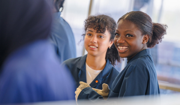 Two students in workshop overalls smile during a practical engineering session in a laboratory environment.