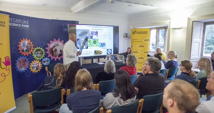 A low resolution image of Professor Ian Thompson stood in front of roller banners and a screen with a PowerPoint presentation on it, presenting to a small room of people.