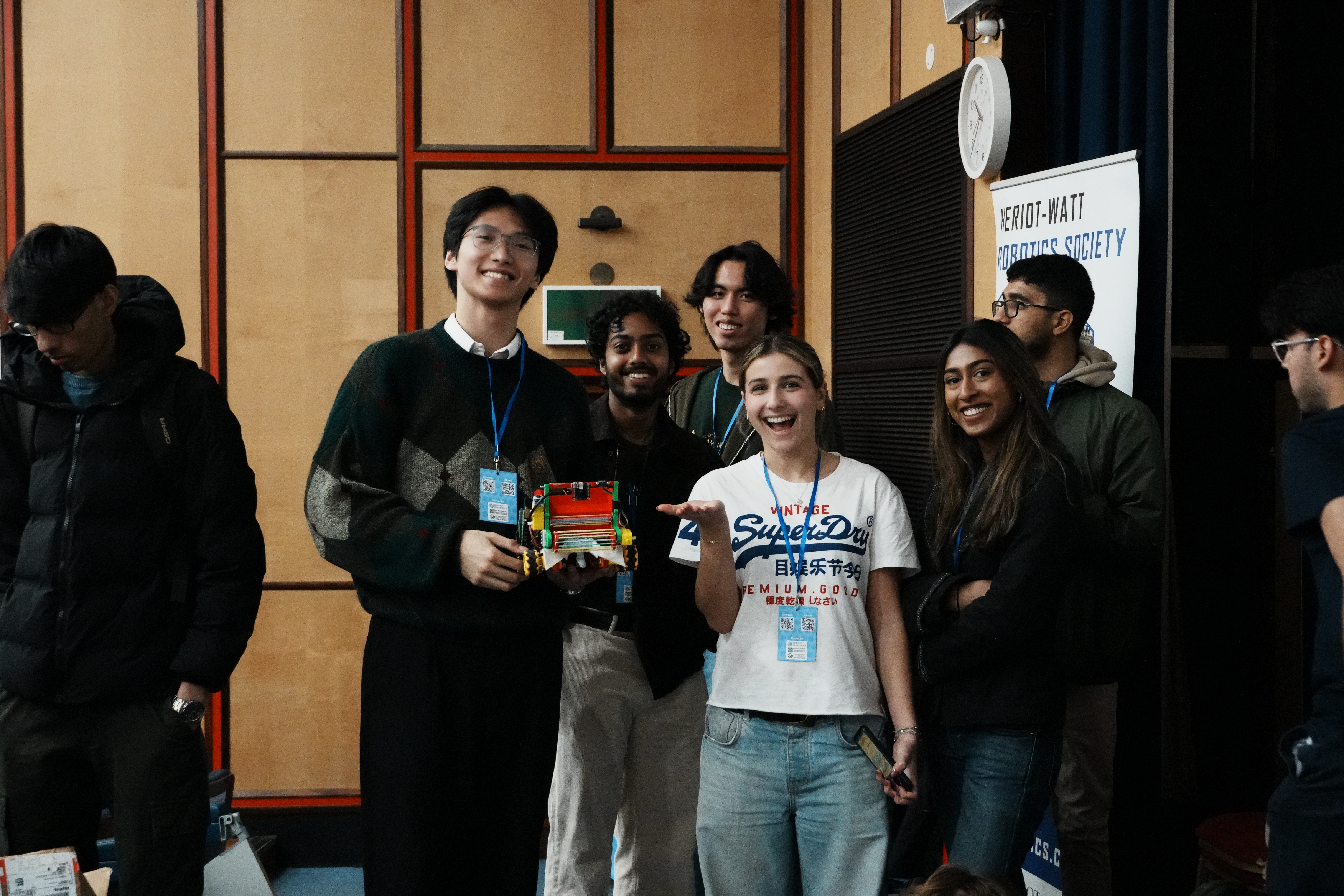 Five students smiling and holding a small robot during a robotics competition event, wearing lanyards.