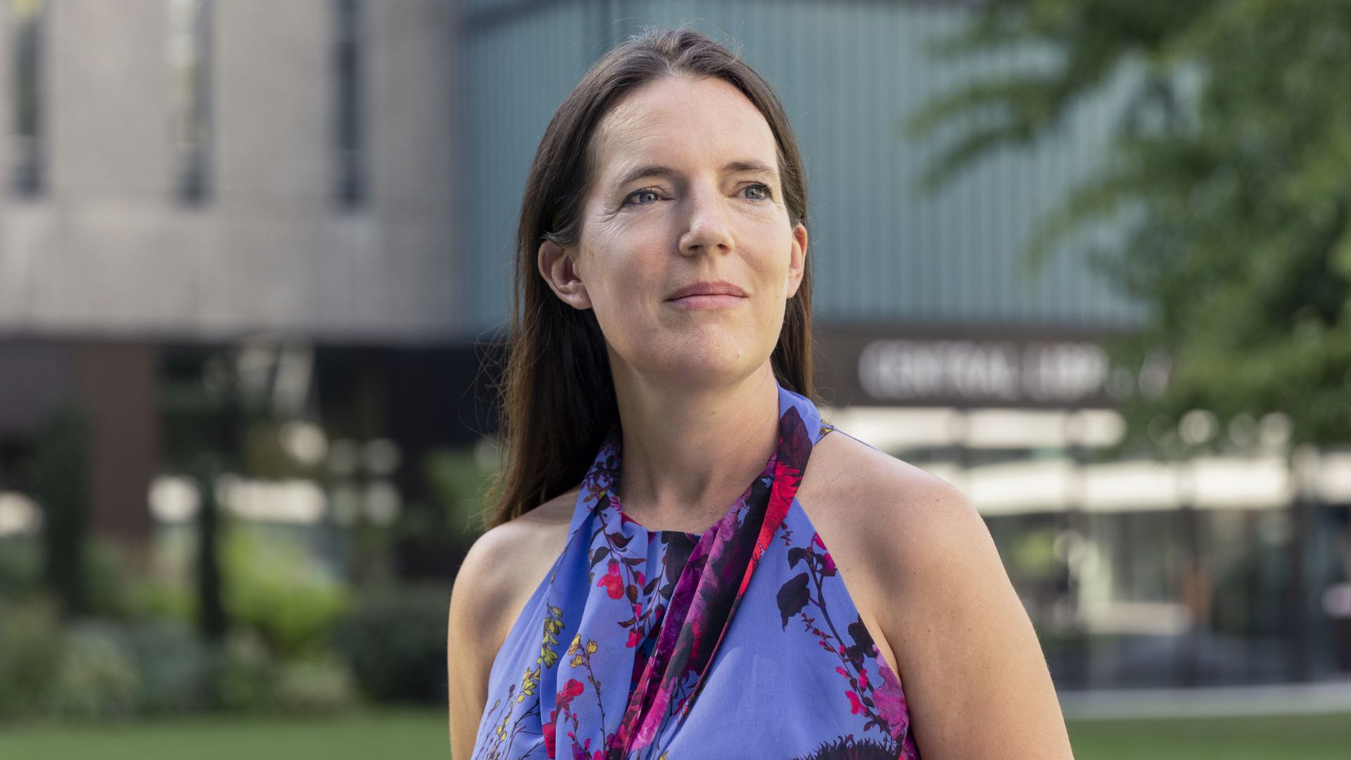 Professor Dame Molly Stevens standing outdoors, looking slightly off-camera. She is wearing a sleeveless floral dress and appears thoughtful. A modern building and greenery are visible in the background.
