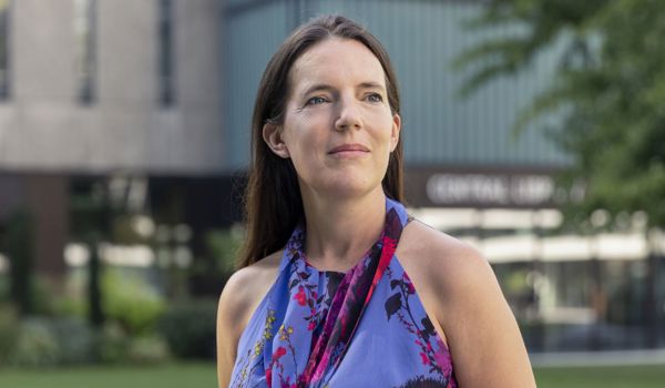 Professor Dame Molly Stevens standing outdoors, looking slightly off-camera. She is wearing a sleeveless floral dress and appears thoughtful. A modern building and greenery are visible in the background.