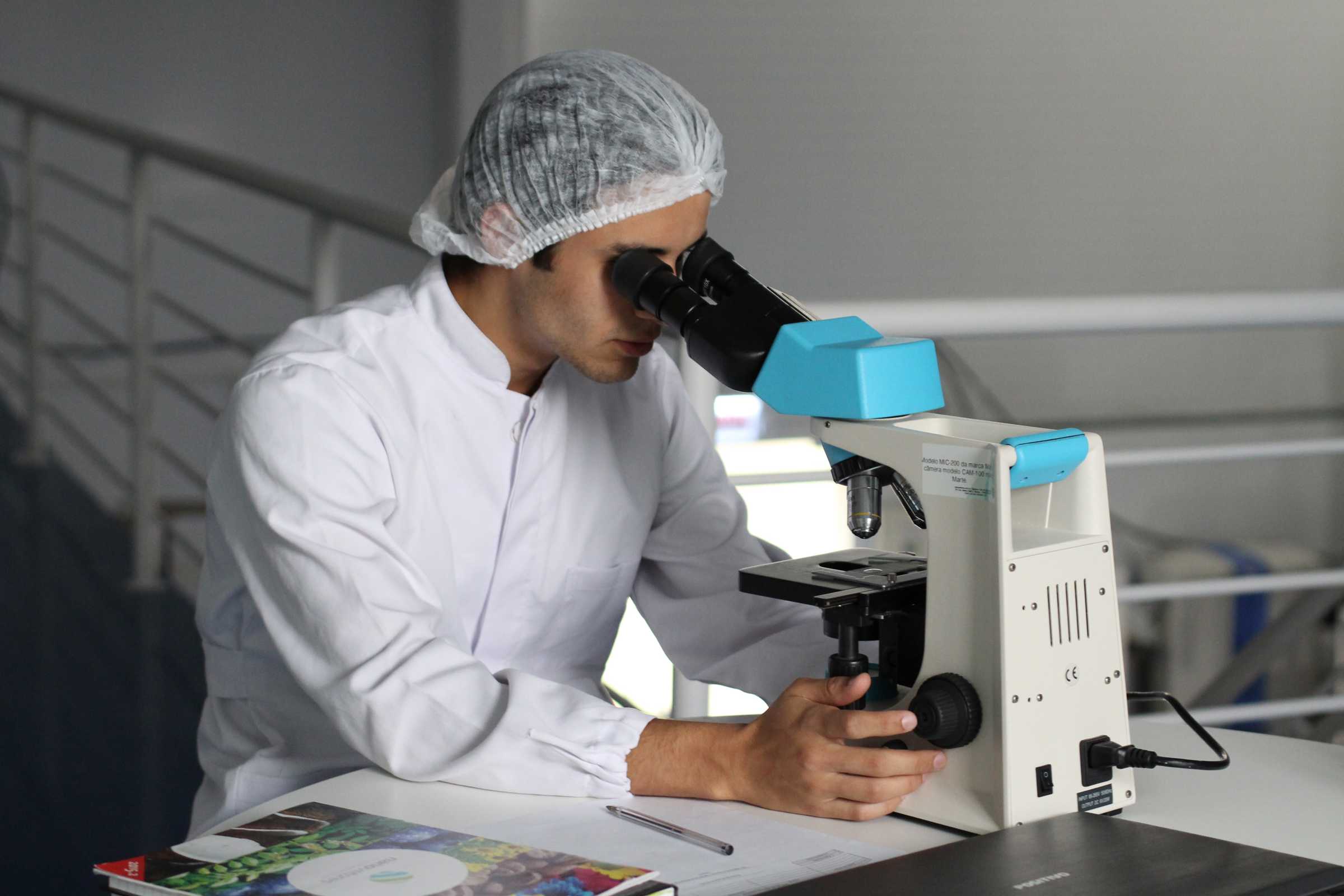 Someone in a white jacket and hairnet looking into a microscope