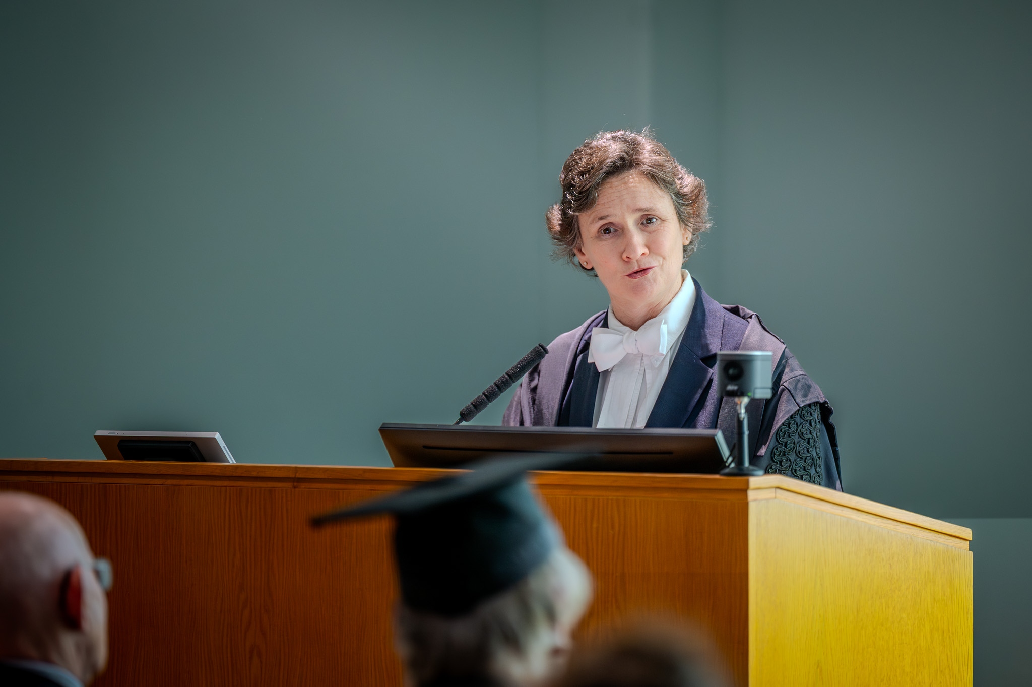 The Vice-Chancellor, Irene Tracey, wearing academic dress, speaks at a lectern during the inaugural John Goodenough Lecture, addressing attendees in a brightly lit hall.
