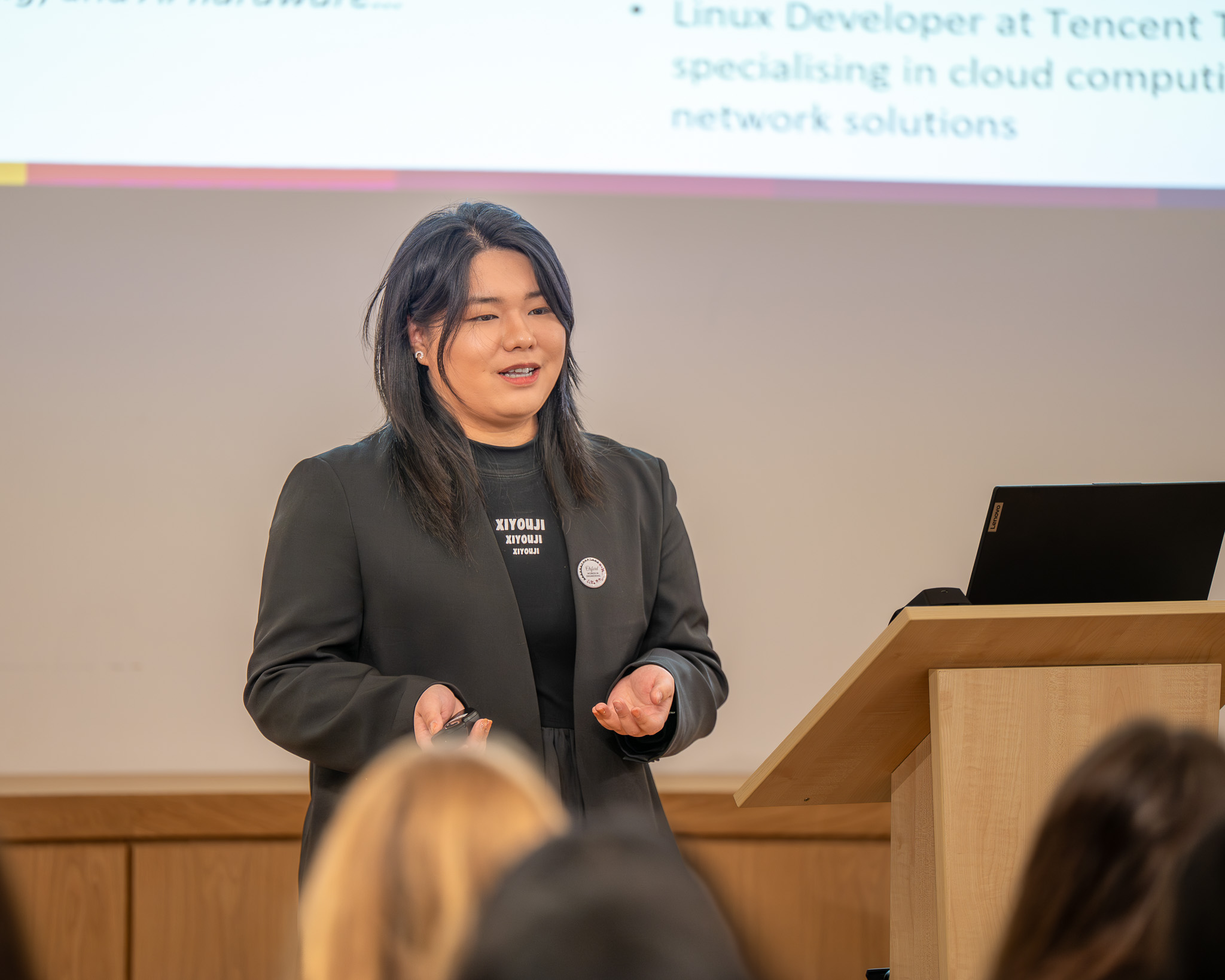 Ziji Chen stands at a lectern speaking to the audience while holding a presentation remote, with part of her slide visible behind her. She is delivering her talk as part of the OxBridge Women in Engineering event.