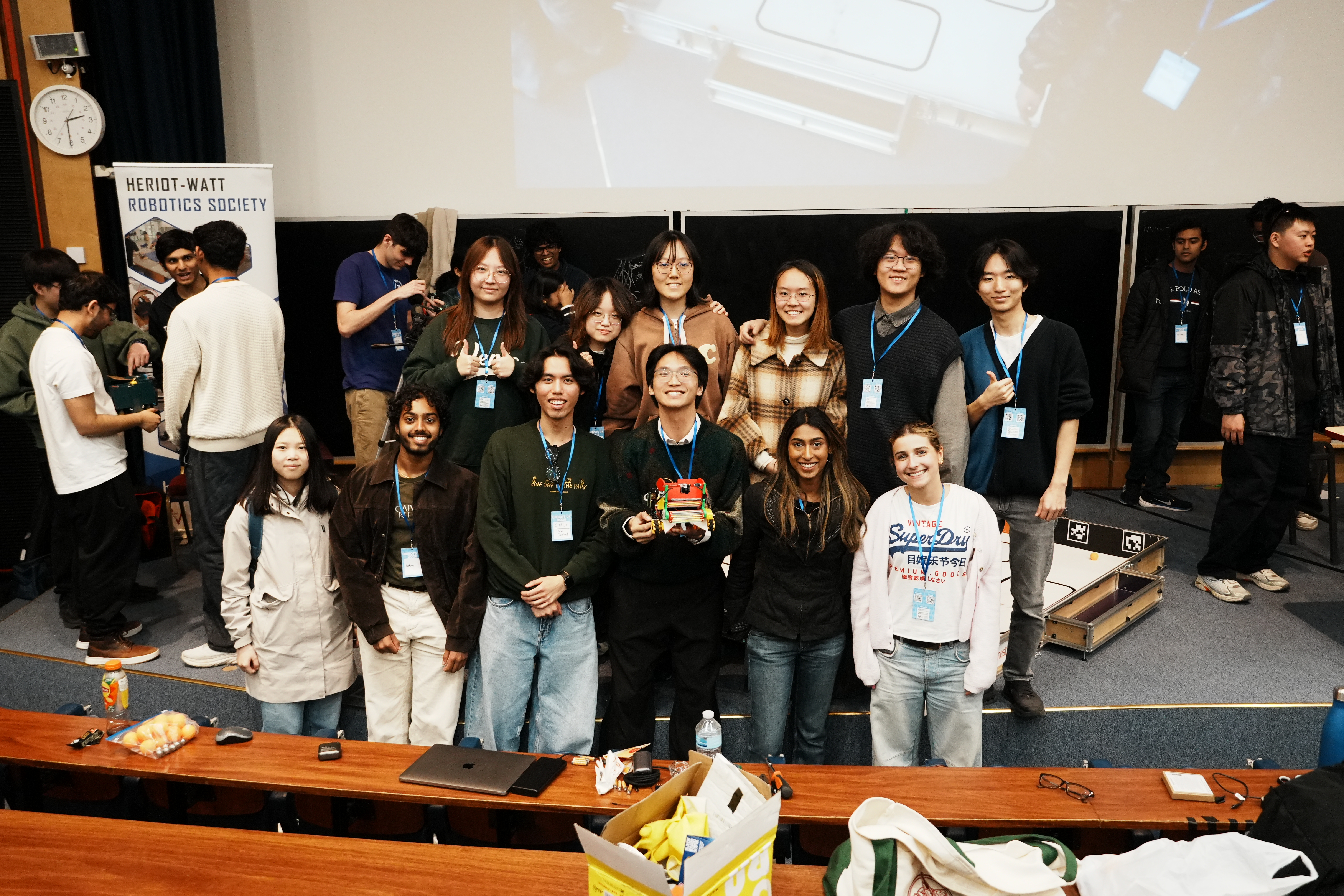 Group of students posing together in a lecture theatre with robotics equipment and competition arena visible in the background.