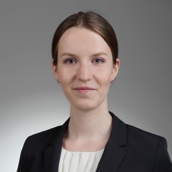 Headshot of DPhil student Sophie Eisele, wearing a black blazer and white top, with a neutral grey background. She is facing the camera and smiling slightly.