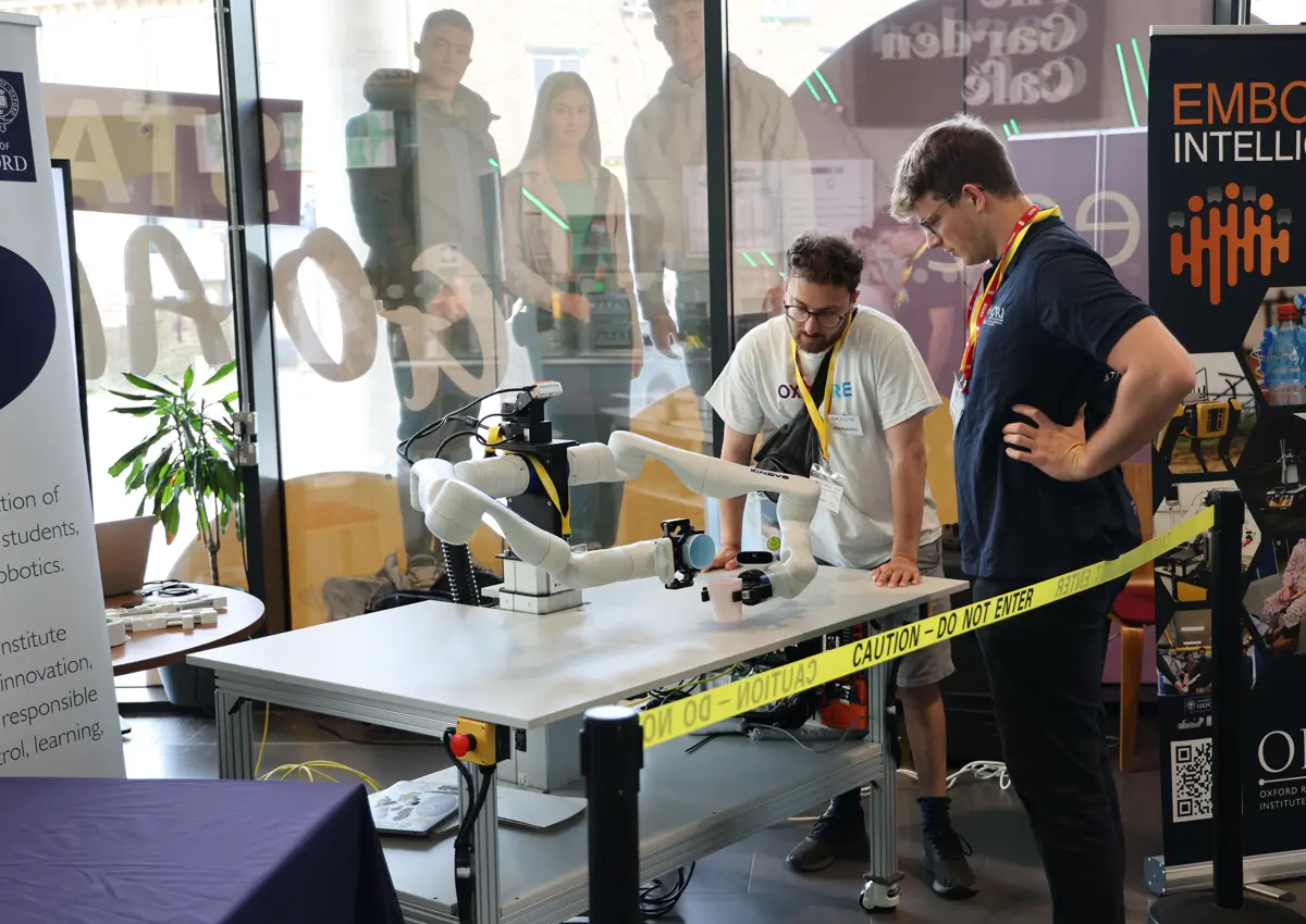 Two Oxford Robotics Institute researchers observe Frank’s interaction with the environment during a demo. The robot is placed on a table surrounded by tech equipment and information stands.