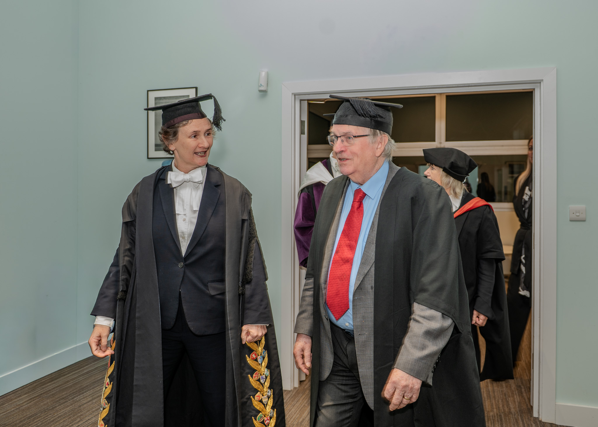 An academic procession enters the room, led by the Bedel in formal robes, followed by senior university figures in academic dress, including the Vice-Chancellor and Professor Stan Whittingham, with additional officials such as the University Assessor and the Master of St Catherine’s College walking behind them.