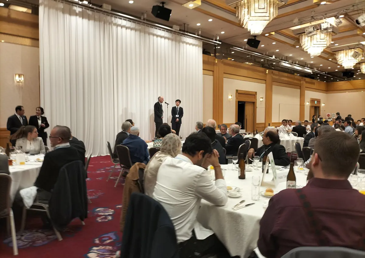 Acceptance Speech: an image of a white man at a mic on stage with an East Asian man. The stage is facing a room of people sat at dining tables.