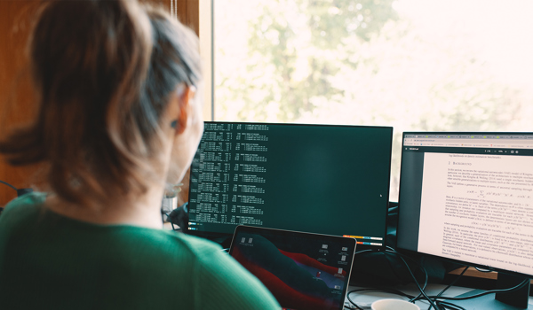 A student with long brown hair tied in a ponytail sits at a desk, working across multiple screens. One monitor displays lines of code, while another shows a scientific research paper. A laptop is also open in front of her. Sunlight streams through a window in the background, casting a warm glow across the workspace.