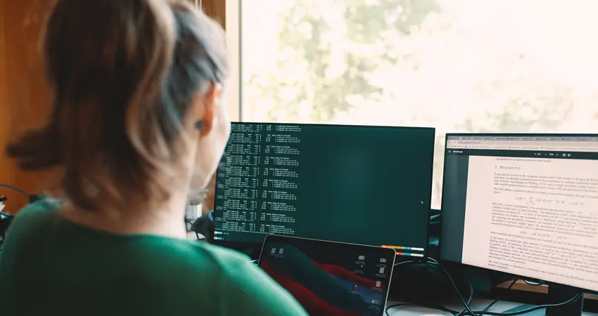 A student with long brown hair tied in a ponytail sits at a desk, working across multiple screens. One monitor displays lines of code, while another shows a scientific research paper. A laptop is also open in front of her. Sunlight streams through a window in the background, casting a warm glow across the workspace.