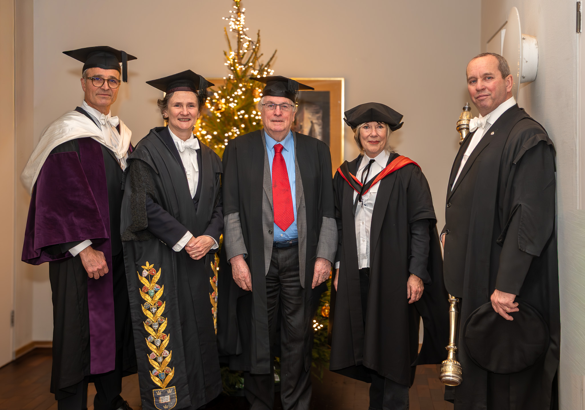A group of five senior university figures in academic dress stand together in a formal setting at St Catherine’s College, positioned in front of a lit Christmas tree. From left to right: the University Assessor, Vice-Chancellor, Professor Stan Whittingham, the Master of St Catherine’s College, and The Bedel holding the ceremonial mace.