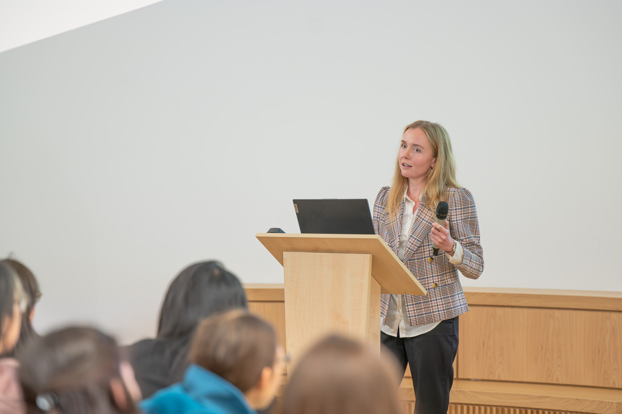 Dr Siân Wilson stands at a lectern holding a microphone while addressing the audience, with a laptop open in front of her. She is delivering her talk on advanced imaging methods as part of the OxBridge Women in Engineering event.