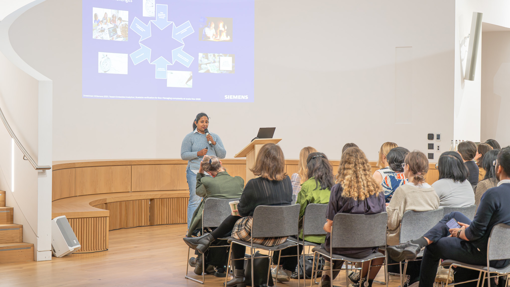 Rasadhi Attale, a Senior Hardware Engineer at Tessent Embedded (Siemens), stands at a lectern addressing the audience, with a presentation slide displaying diagrams behind her. She is delivering her talk as part of the OxBridge Women in Engineering event.