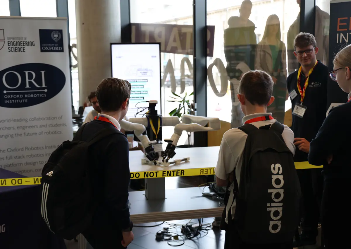An Oxford Robotics Institute researcher stands next to Frank the robot at a demonstration booth, engaging students at the Oxplore Festival in Bradford. A branded banner and STEM posters are visible in the background.