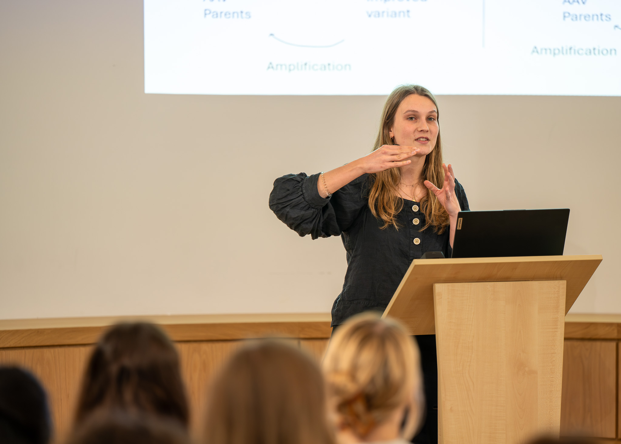 Marina Luchner stands at a lectern addressing the audience, gesturing with her hands as she explains her research, with part of her presentation slide visible behind her. She is speaking as part of the OxBridge Women in Engineering event.