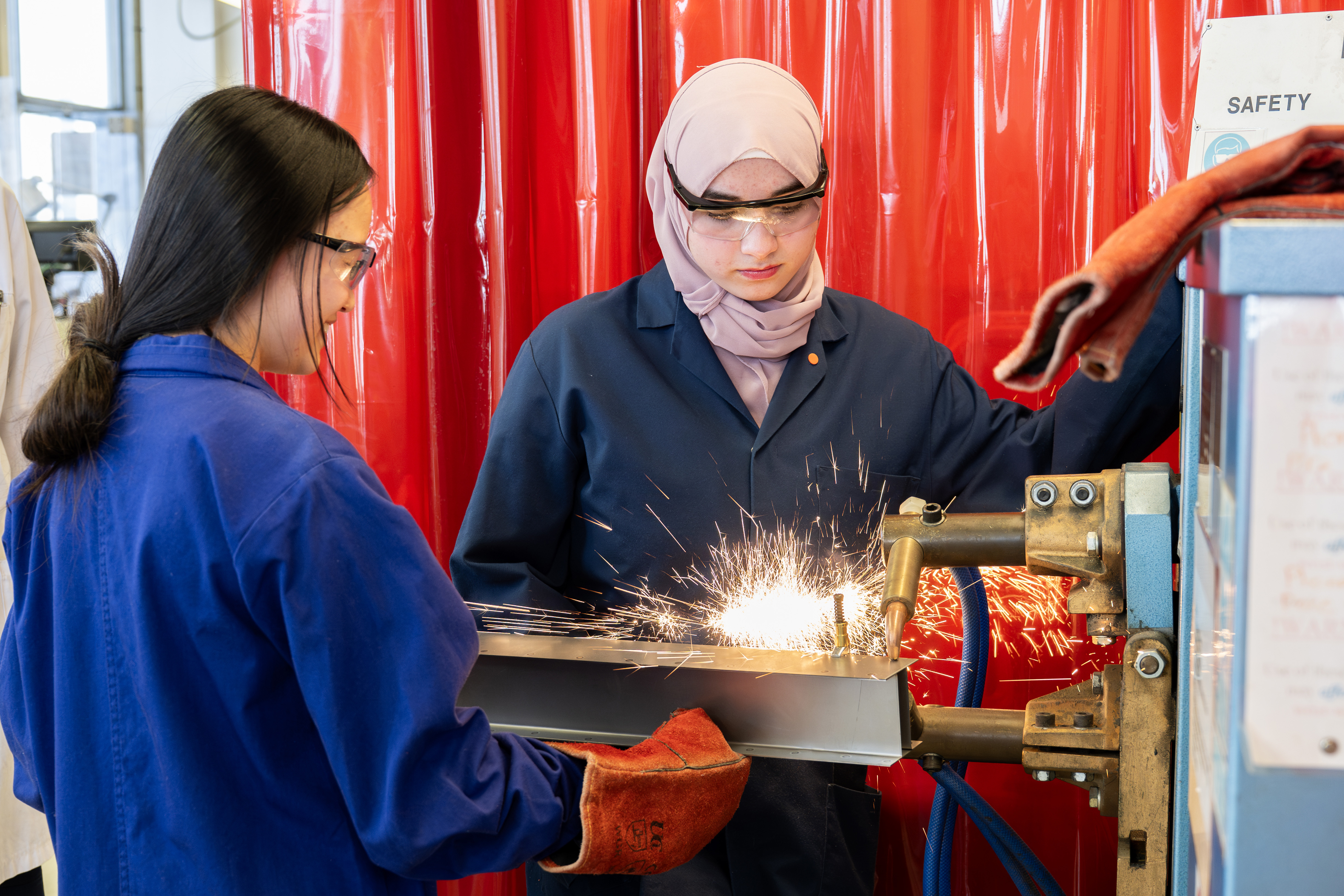 Two undergraduate engineering students wearing protective lab coats, gloves, and safety goggles operate welding equipment in the University of Oxford’s Design, Build, and Test Lab. Bright sparks fly as they work on a metal beam, with red safety curtains in the background.
